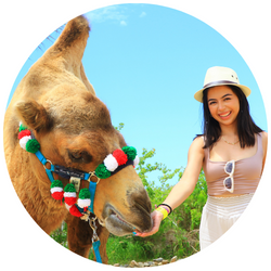 photo of Geraldine Lopez, a UCR student with dark hair, wearing a hat, smiling at the camera and feeding a camel