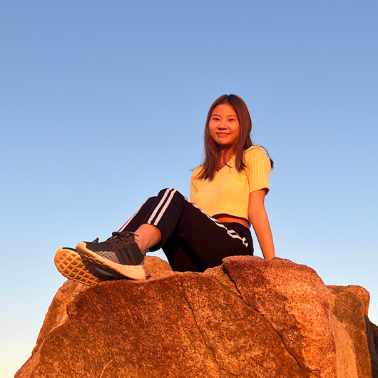 Female, early 20s, dark hair, sitting on a large rock at sunset