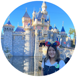 Photo of Janet Maringka, a UCR student with brown hair who is smiling at the camera and standing in front of a castle at Disneyland
