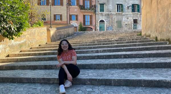 Female, early 20s, with dark hair, sitting on stone steps in front of old stone buildings in Italy